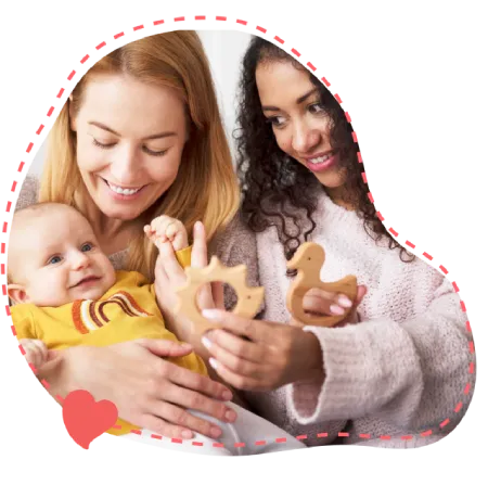 Two smiling women hold wooden teething toys with a baby in a yellow onesie